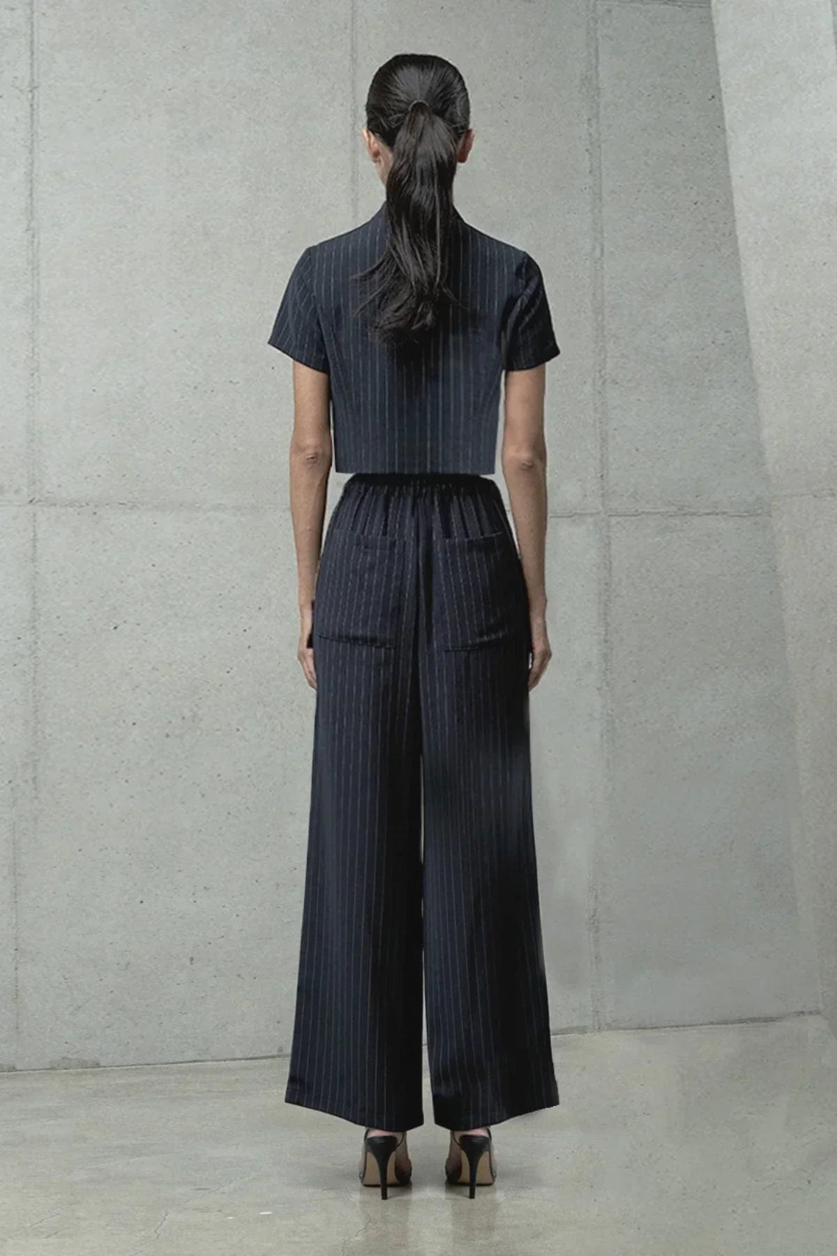 Woman with long ponytail wearing black pinstripe short-sleeve crop top and wide-leg pants in minimal concrete setting