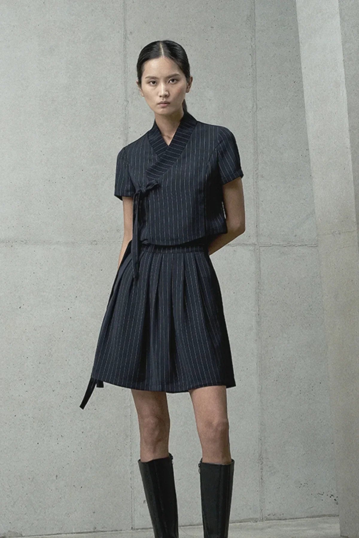Model in black pinstripe short-sleeve top and pleated skirt with black knee-high boots against concrete wall