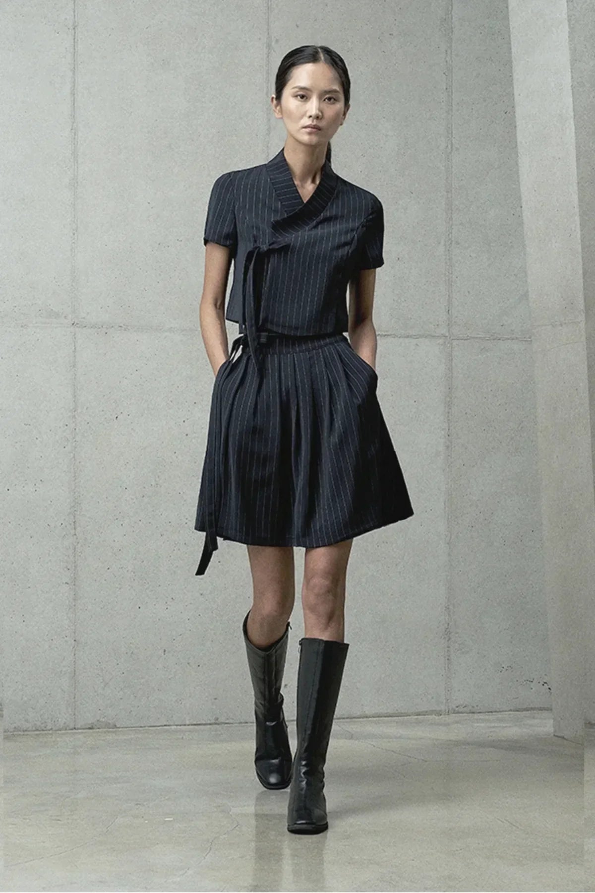 Model wearing a black pinstripe short-sleeve jeogori top with matching pleated skirt and black knee-high boots in studio