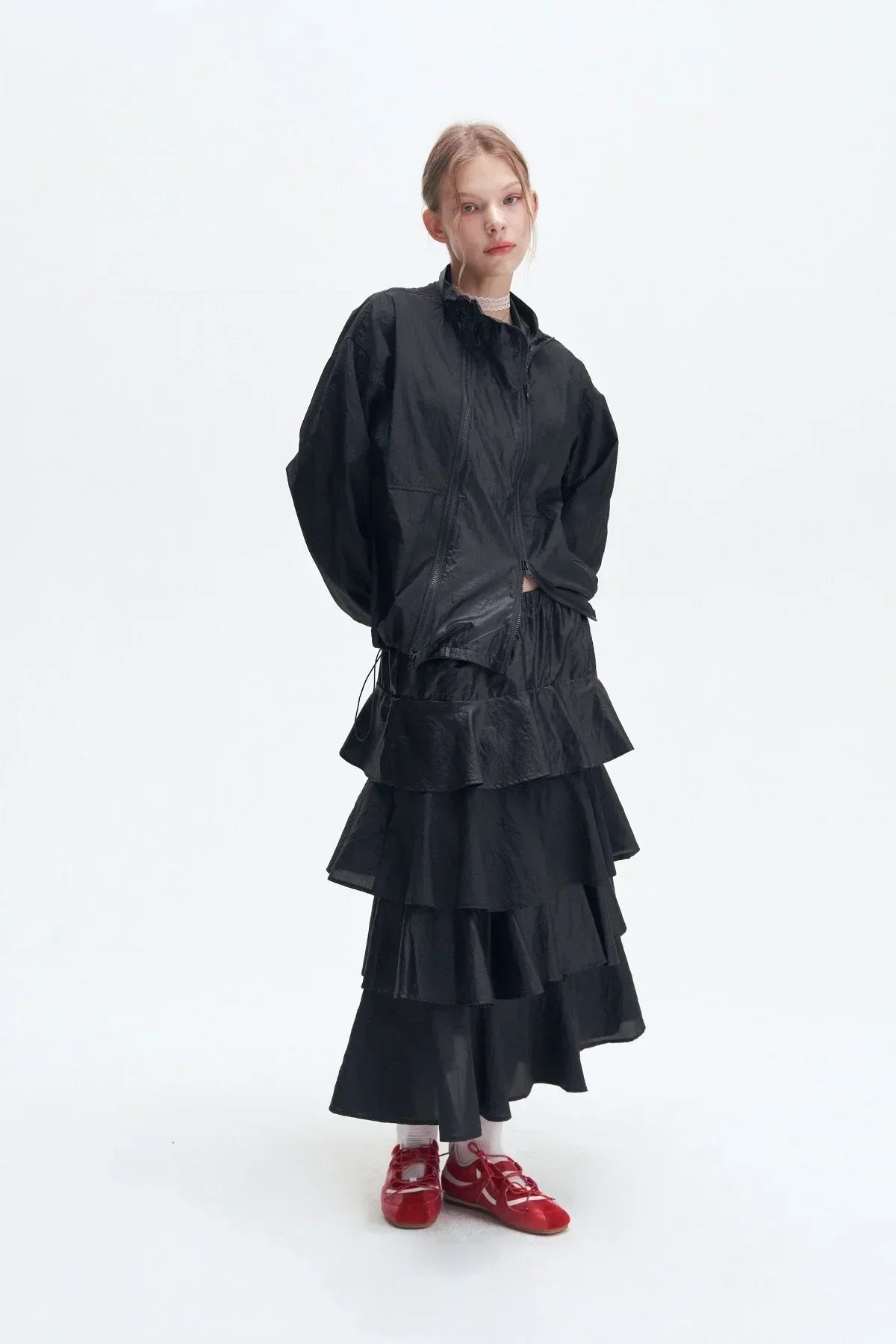 Model wearing a black double zipper windbreaker jacket and layered ruffle black skirt with red shoes on white background