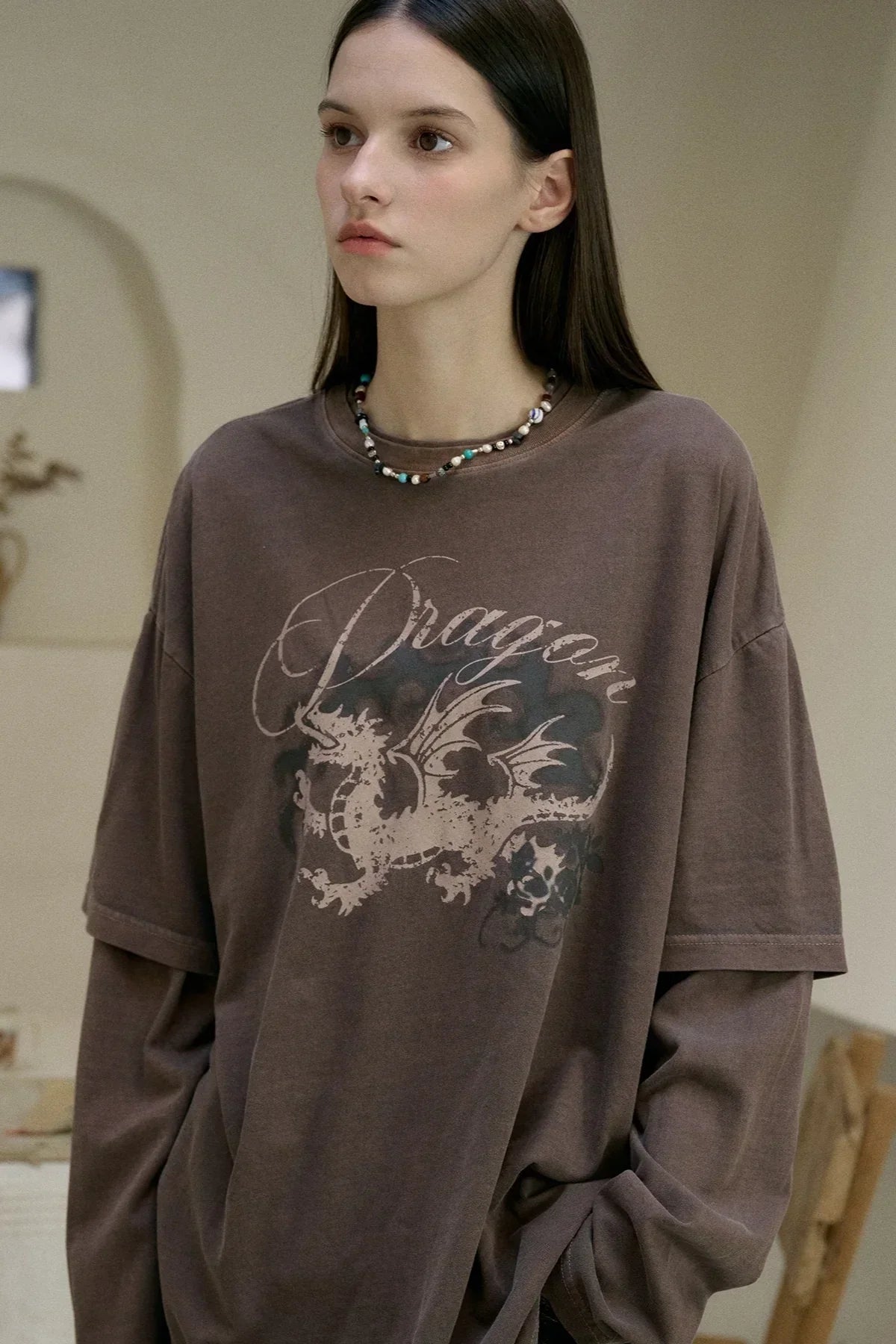 Woman wearing brown layered long sleeve shirt with dragon graphic and beaded necklace in neutral room