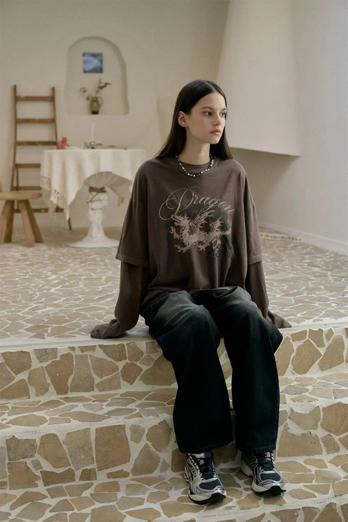 Young woman in brown dragon print layered long sleeve shirt and black pants sitting on mosaic tile steps in minimalist room