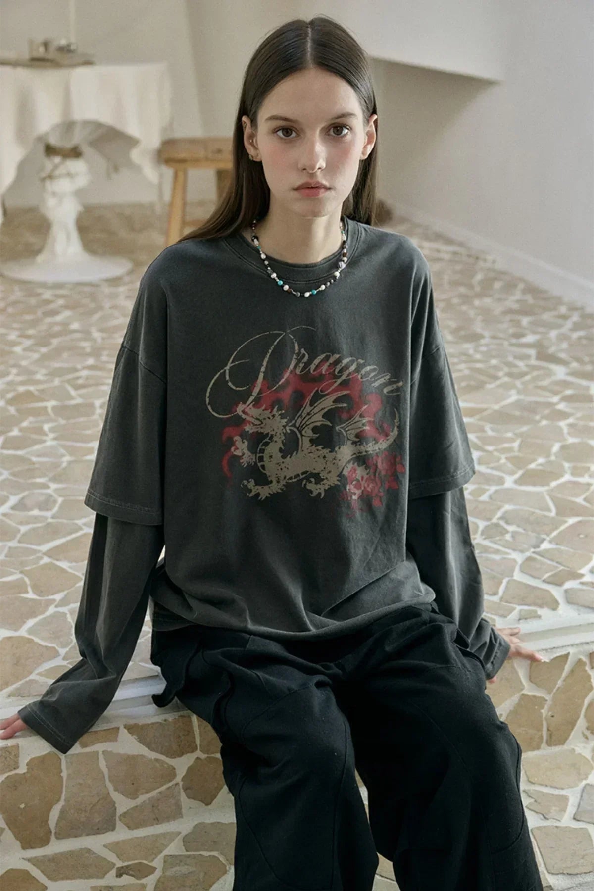 Young woman sitting on mosaic tile floor wearing smoke black layered long sleeve shirt with dragon print