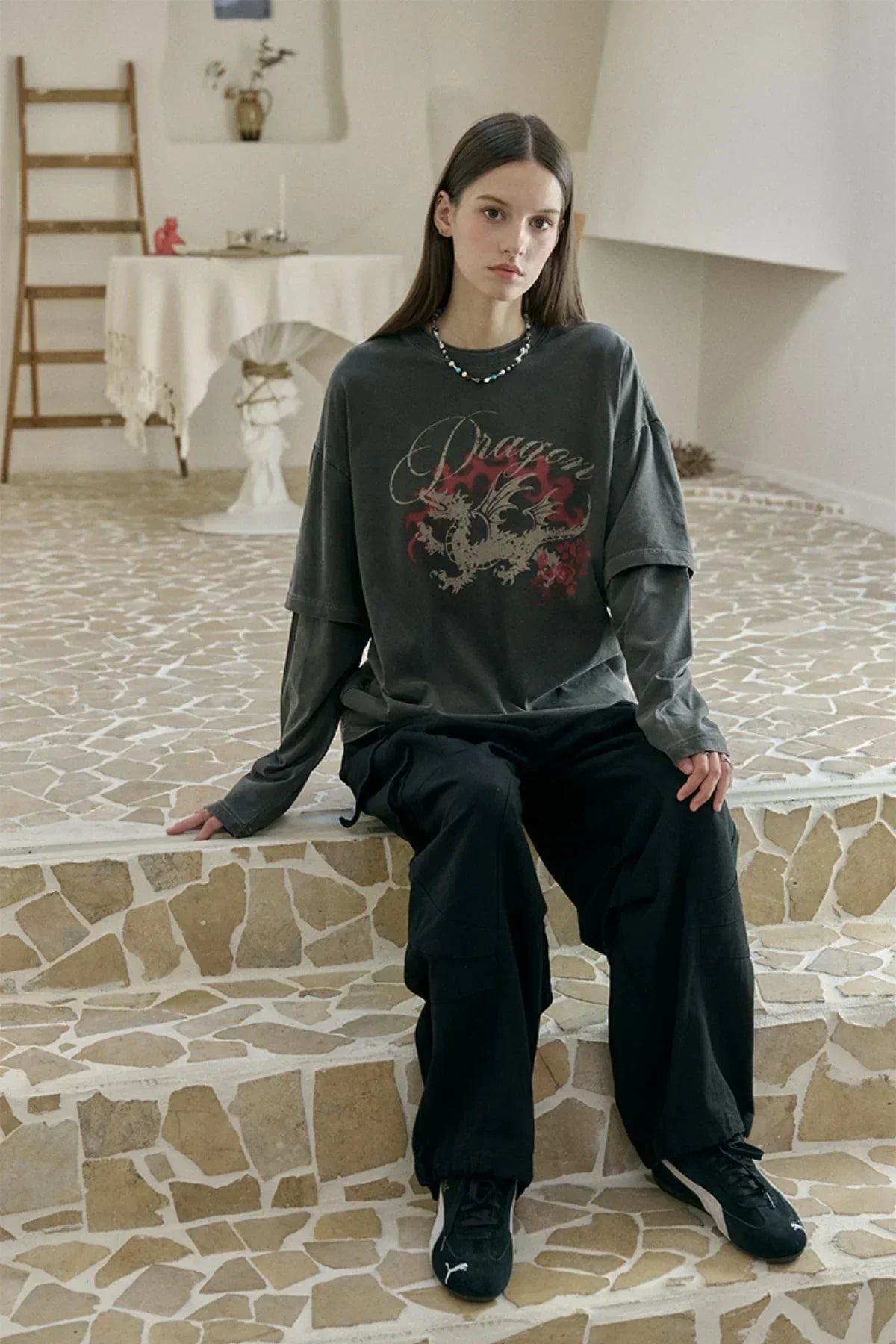 Young woman in layered dragon graphic long sleeve shirt and black cargo pants sitting on stone steps indoors
