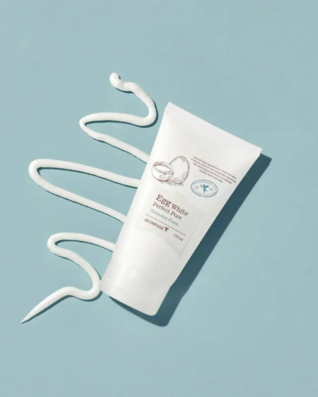 Skinfood Egg White Perfect Pore Cleansing Foam tube on blue surface with creamy texture swirl
