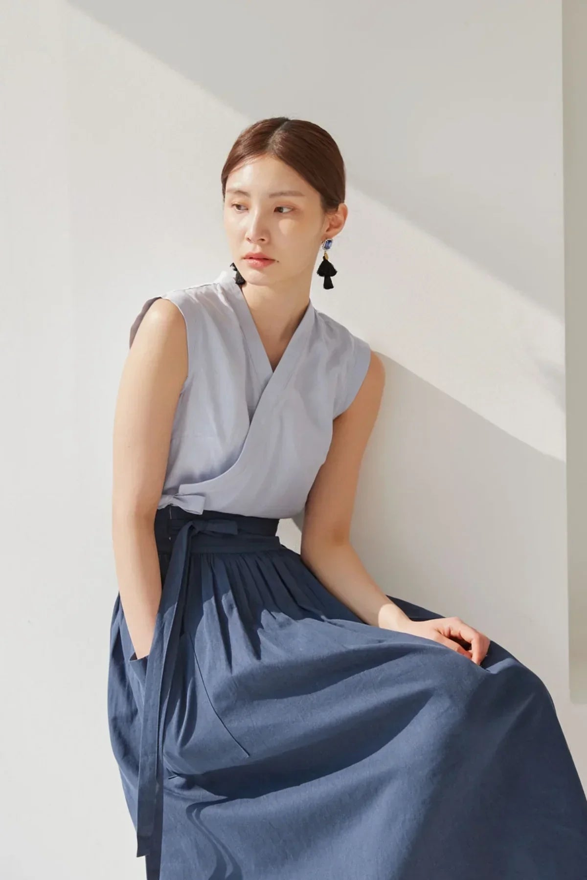 Woman sitting in natural light wearing sleeveless light blue wrap top and navy blue pleated skirt with pockets