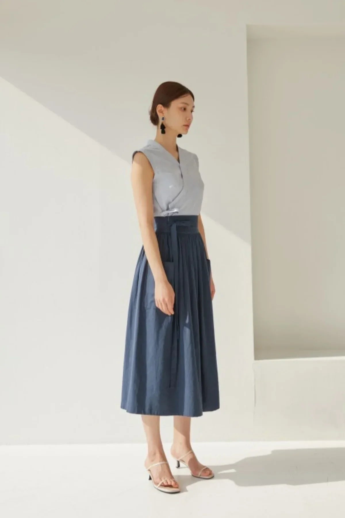 Woman wearing sleeveless gray top and navy blue midi skirt with pockets in bright minimalist setting