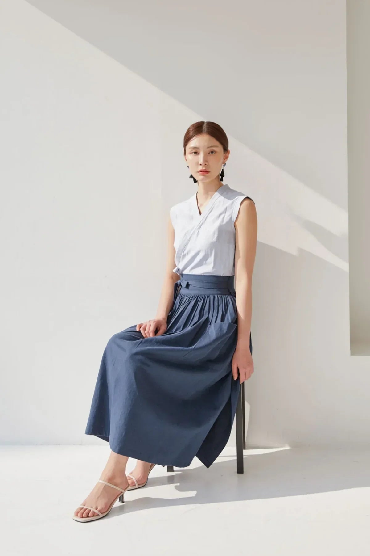 Woman sitting on chair wearing sleeveless white top and navy blue midi skirt in bright minimalist studio