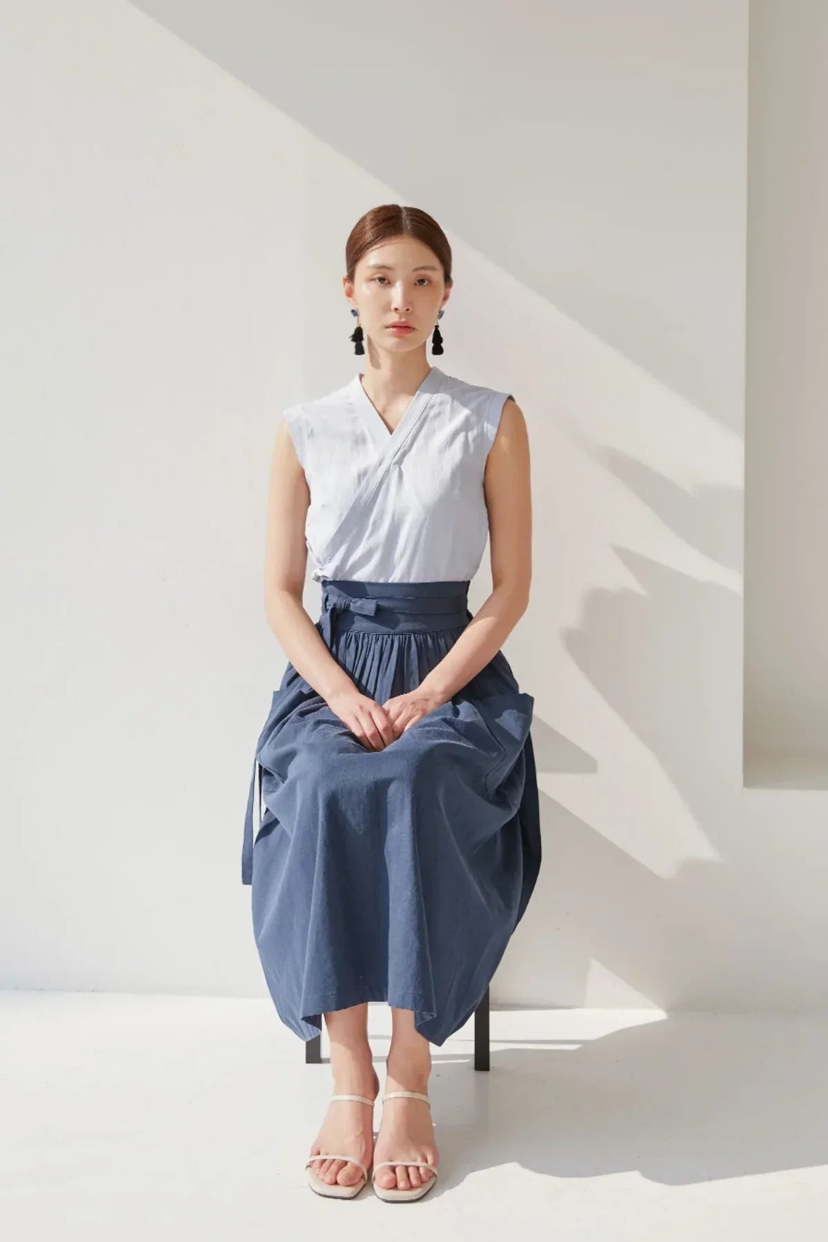 Woman sitting on a chair wearing a sleeveless white top, navy blue skirt, and white sandals in bright studio