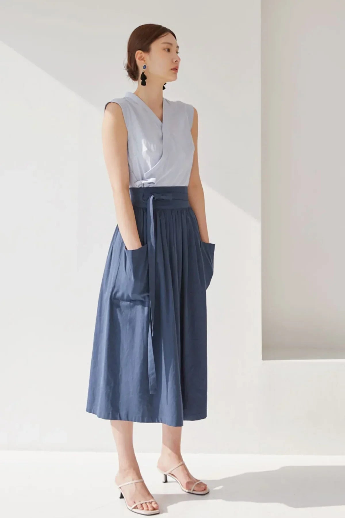 Woman in sleeveless light blue top and navy blue midi skirt with pockets, posing in minimalist white studio