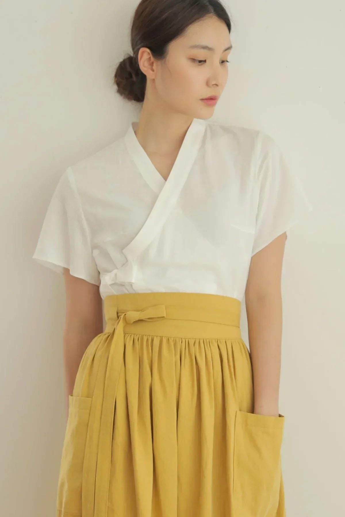 Young woman wearing white wrap blouse and mustard yellow skirt with pockets, standing against light wall