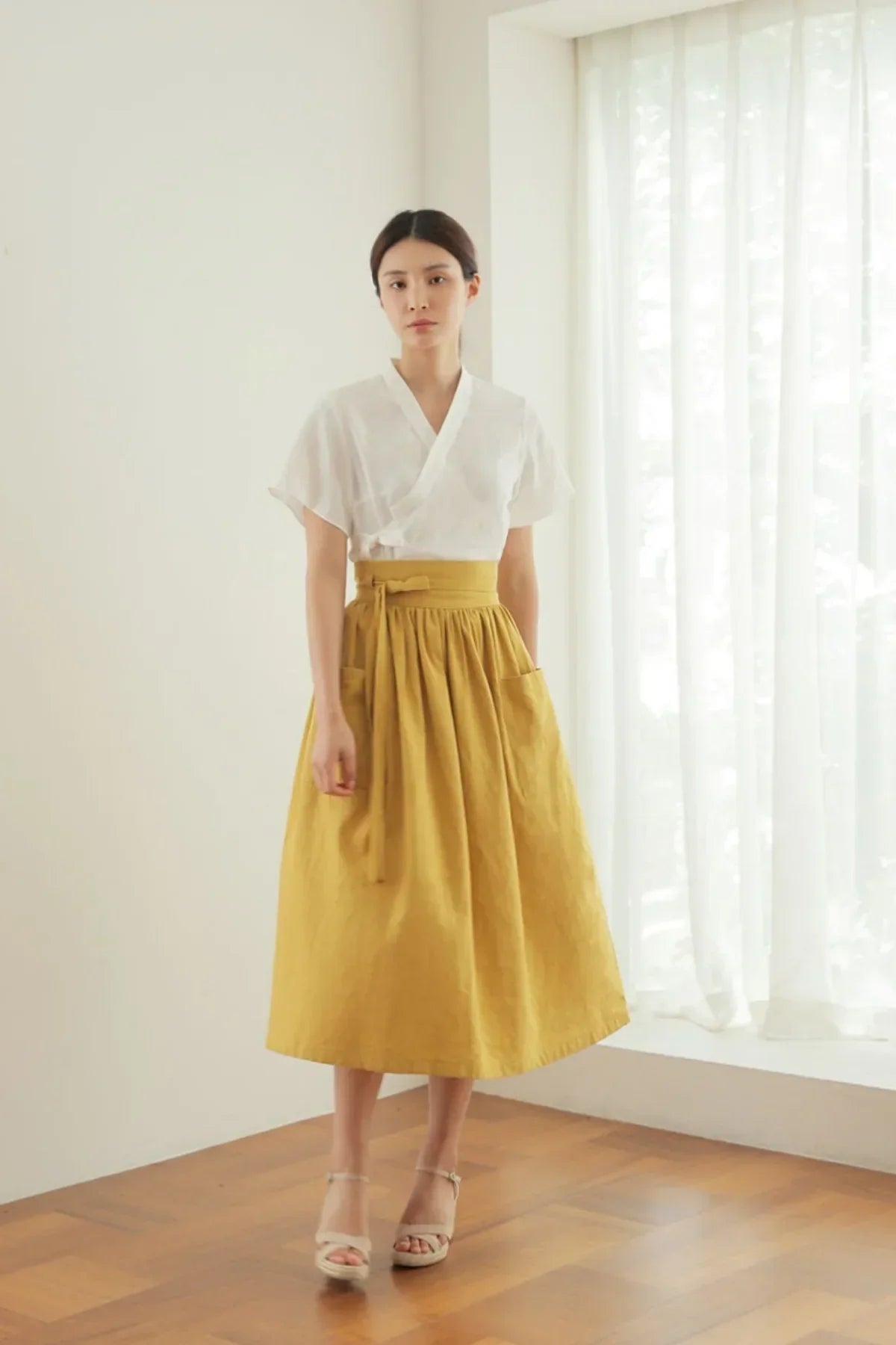 Woman wearing mustard yellow high-waisted skirt with pockets and white wrap top in minimalist room