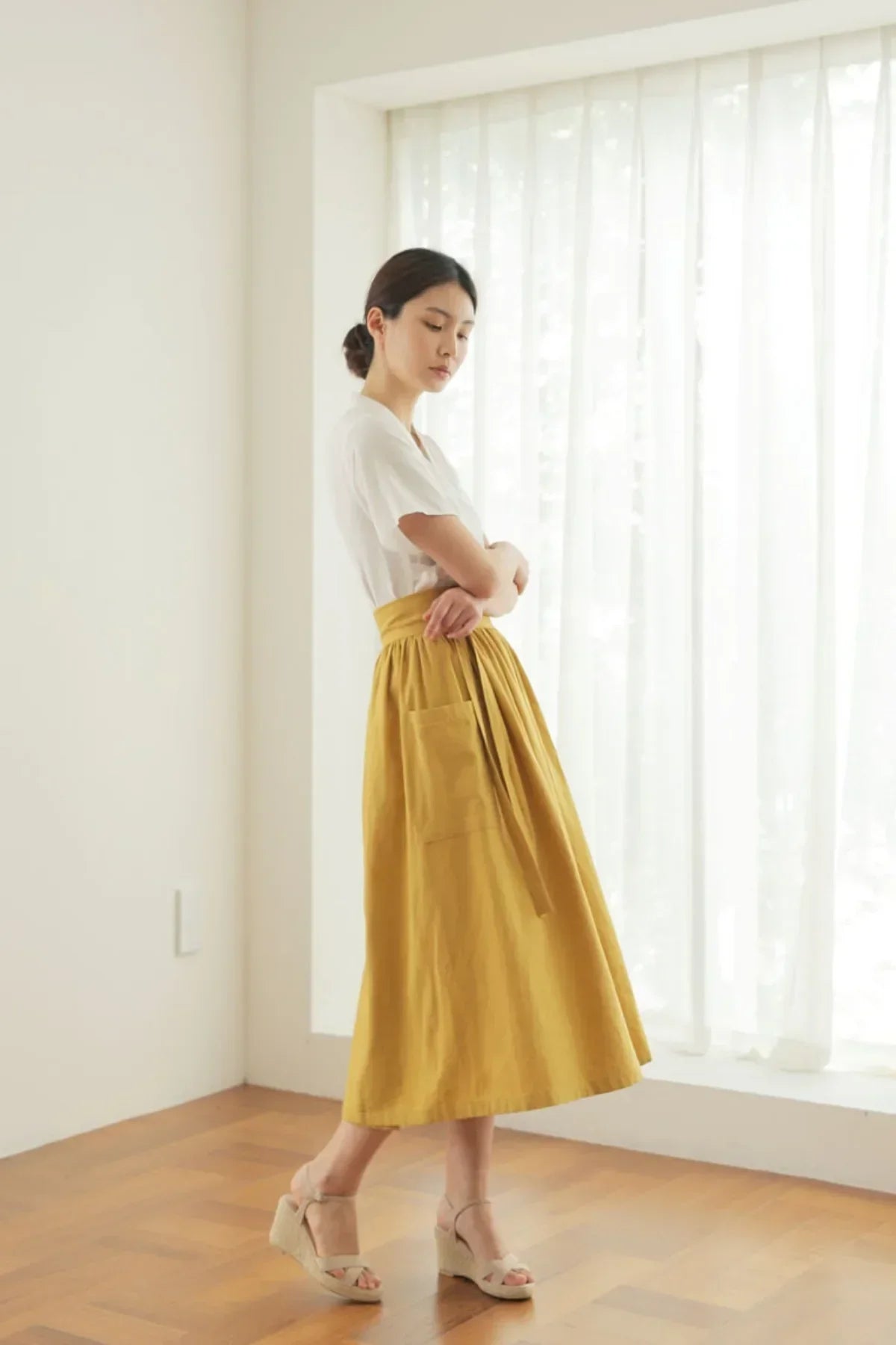 Woman in white blouse and mustard midi skirt with pocket, standing near sheer curtains in bright room