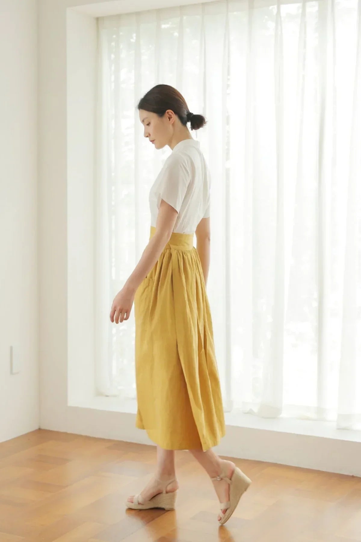 Woman wearing mustard midi skirt and white blouse walking on wooden floor by sheer white curtains