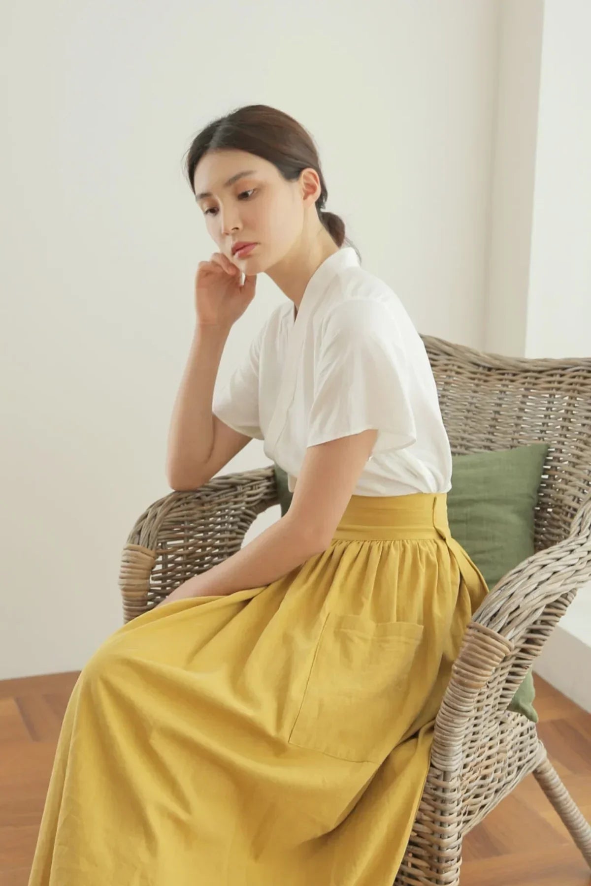 Woman wearing a mustard yellow skirt and white blouse sitting on a wicker chair with a green cushion