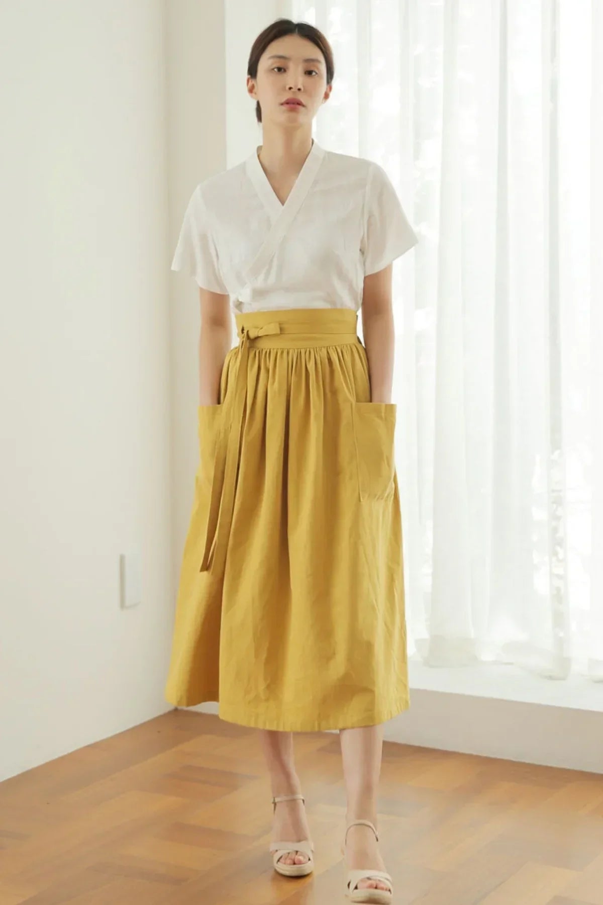 Woman wearing mustard yellow high-waisted skirt with oversized pockets and white short-sleeve top indoors