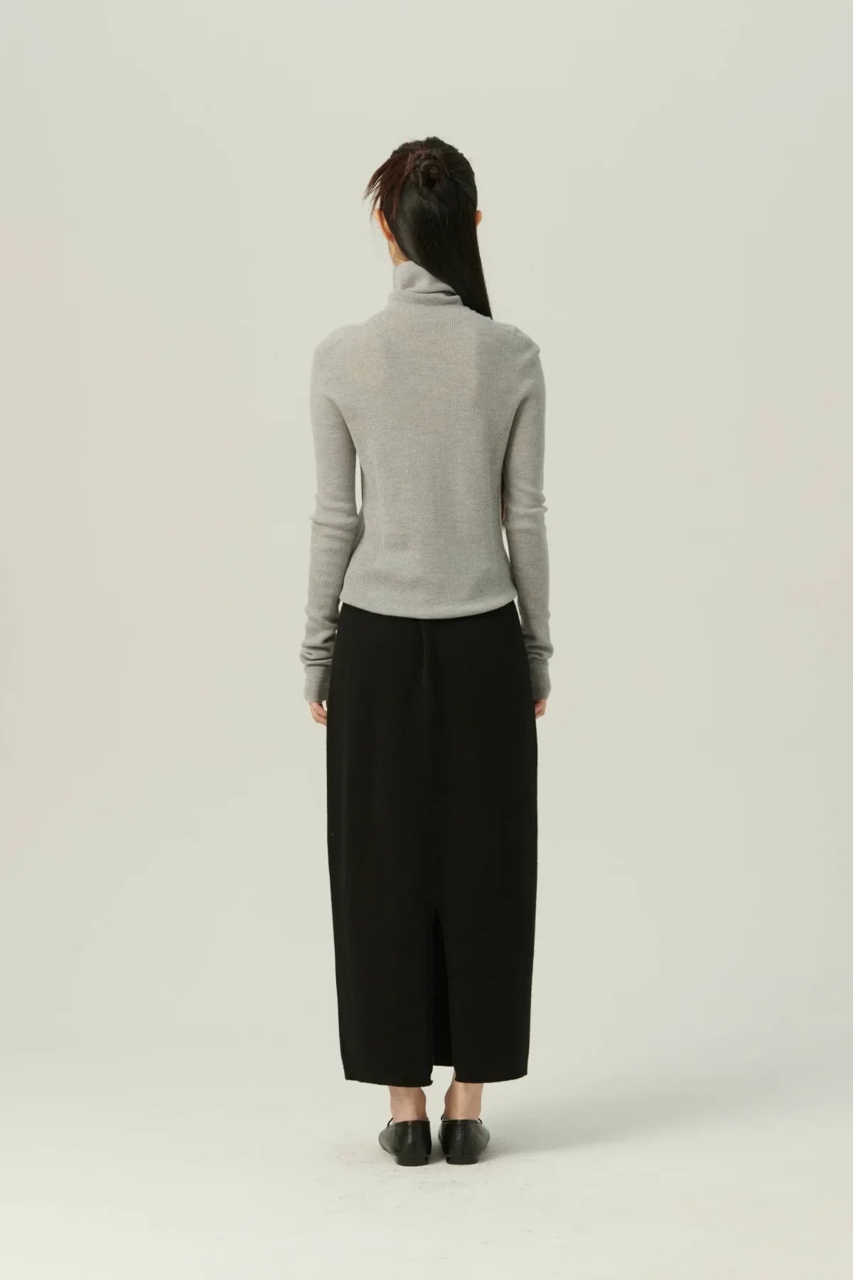 Back view of woman wearing gray fine rib wool turtleneck and long black skirt with black shoes