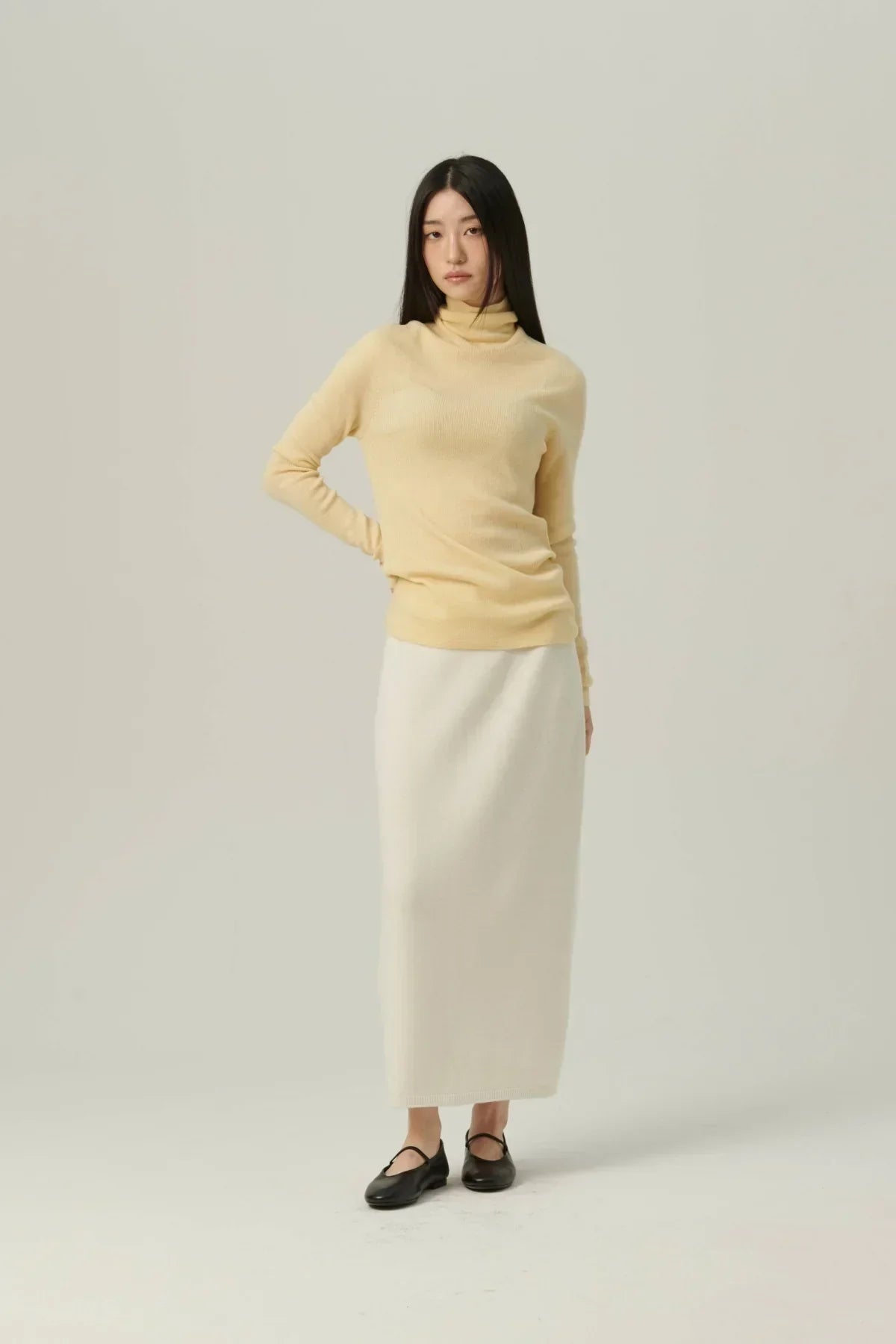 Woman wearing yellow fine rib wool turtleneck sweater and long white skirt with black flats on neutral background