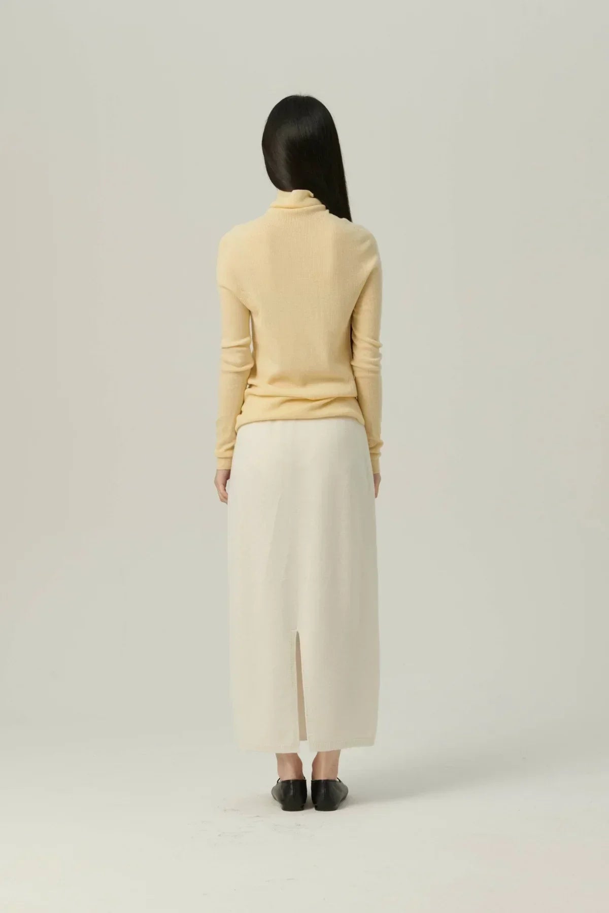 Back view of woman in yellow wool turtleneck and white midi skirt with slit, black flats on plain background