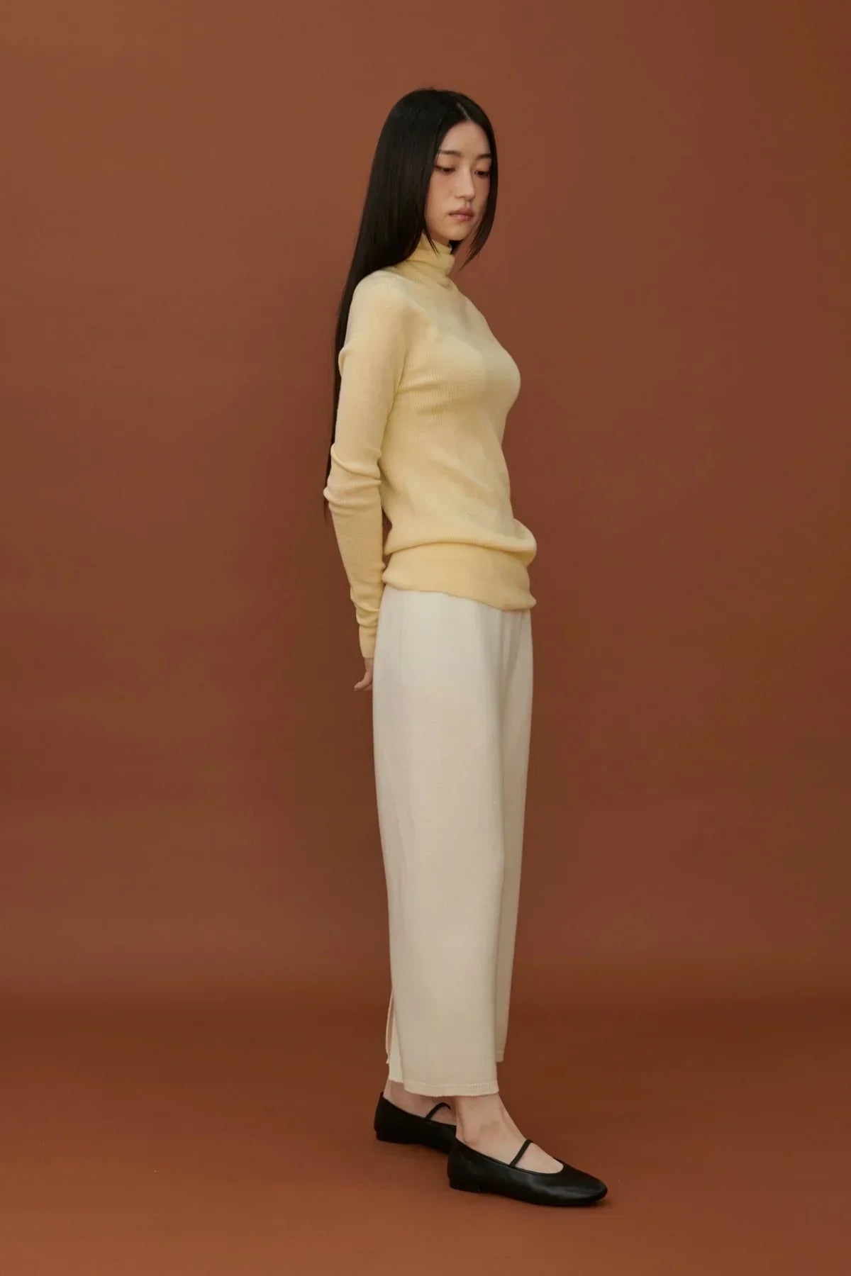 Side view of woman in yellow fine rib wool turtleneck and white wide pants with black flats on brown background