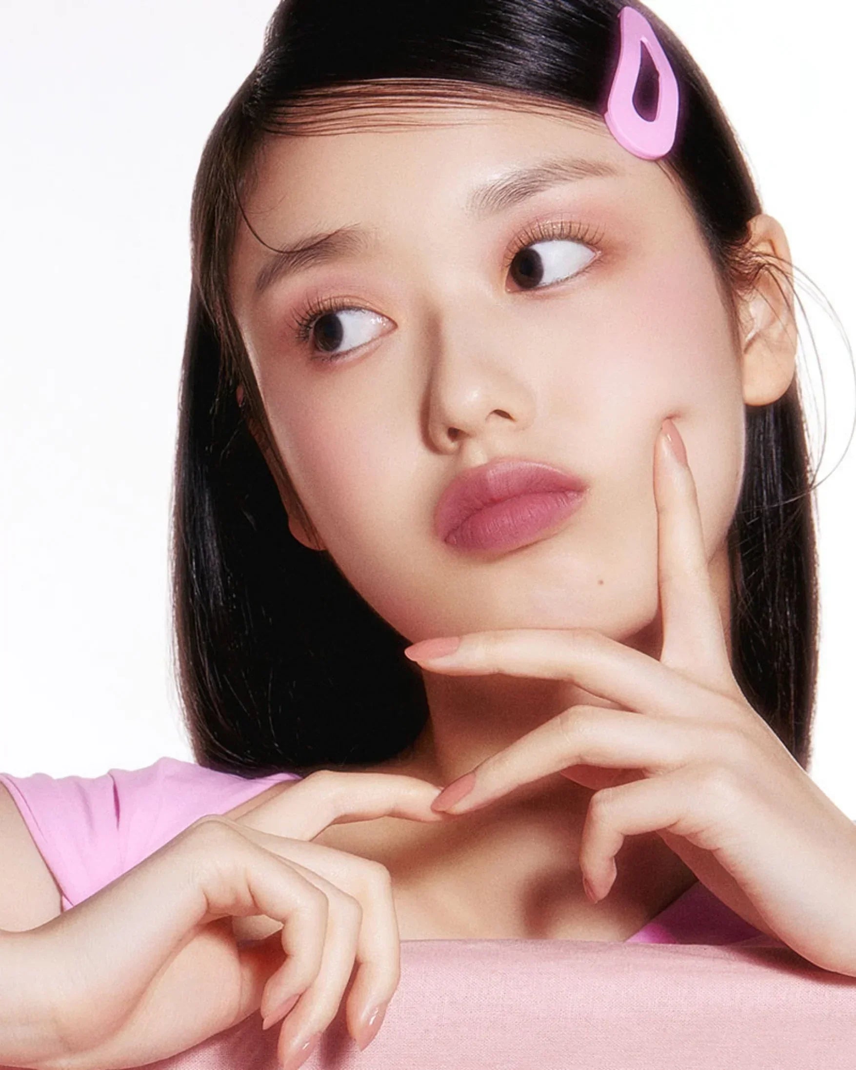 Young woman with smooth skin, pink lipstick, pink top, and pink hair clip posing with hands near face