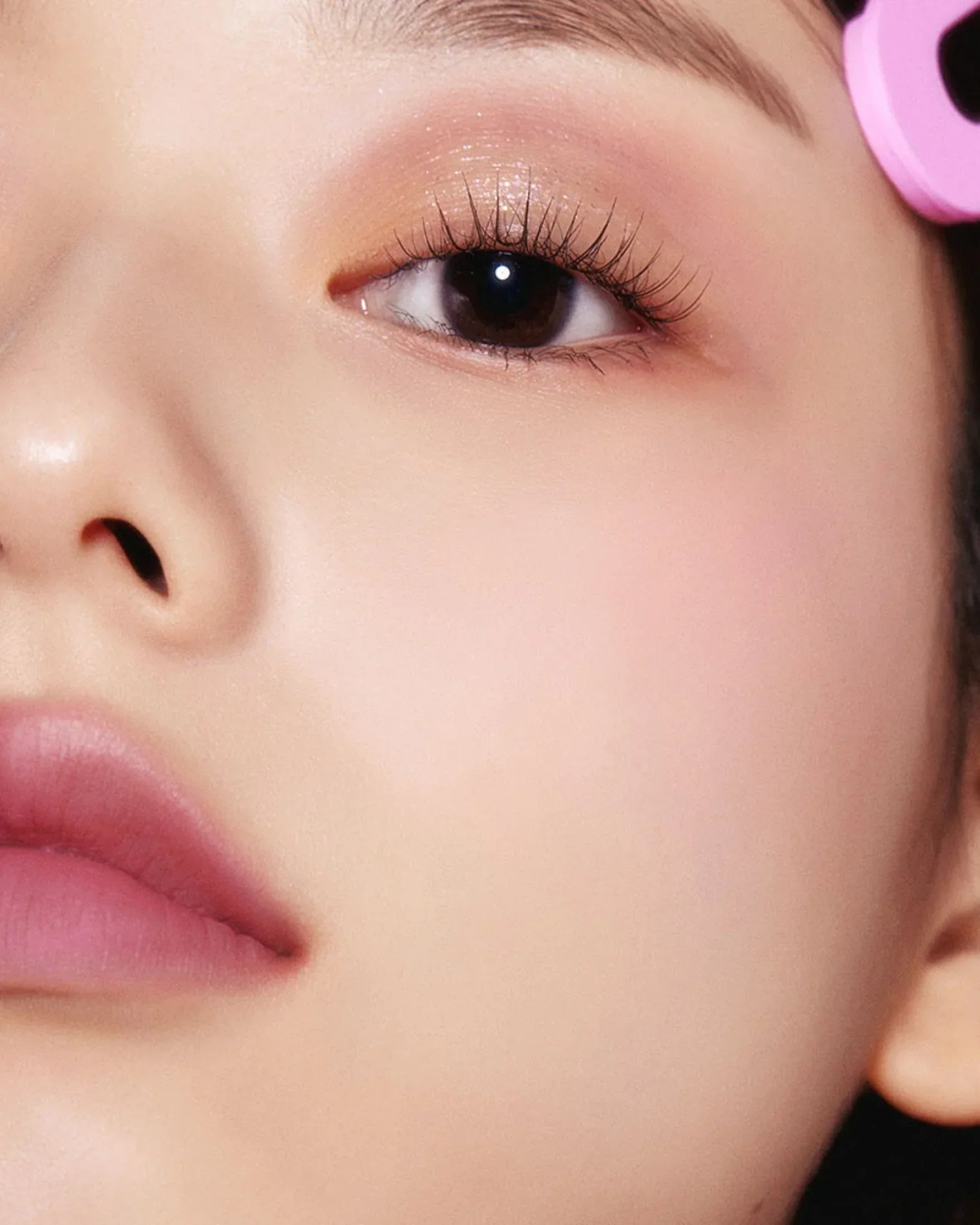 Close-up of a woman's eye with long lashes and soft pink makeup, natural skin, and a pink hair roller