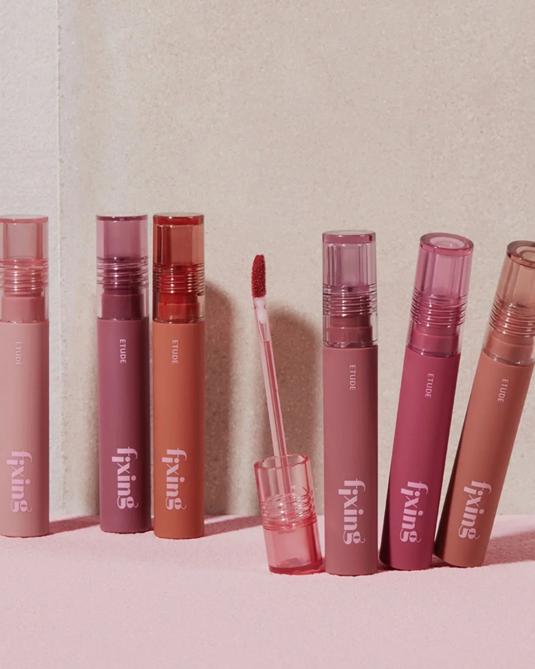 Row of Etude Fixing tint lipsticks in various muted pink and brown shades on pink surface