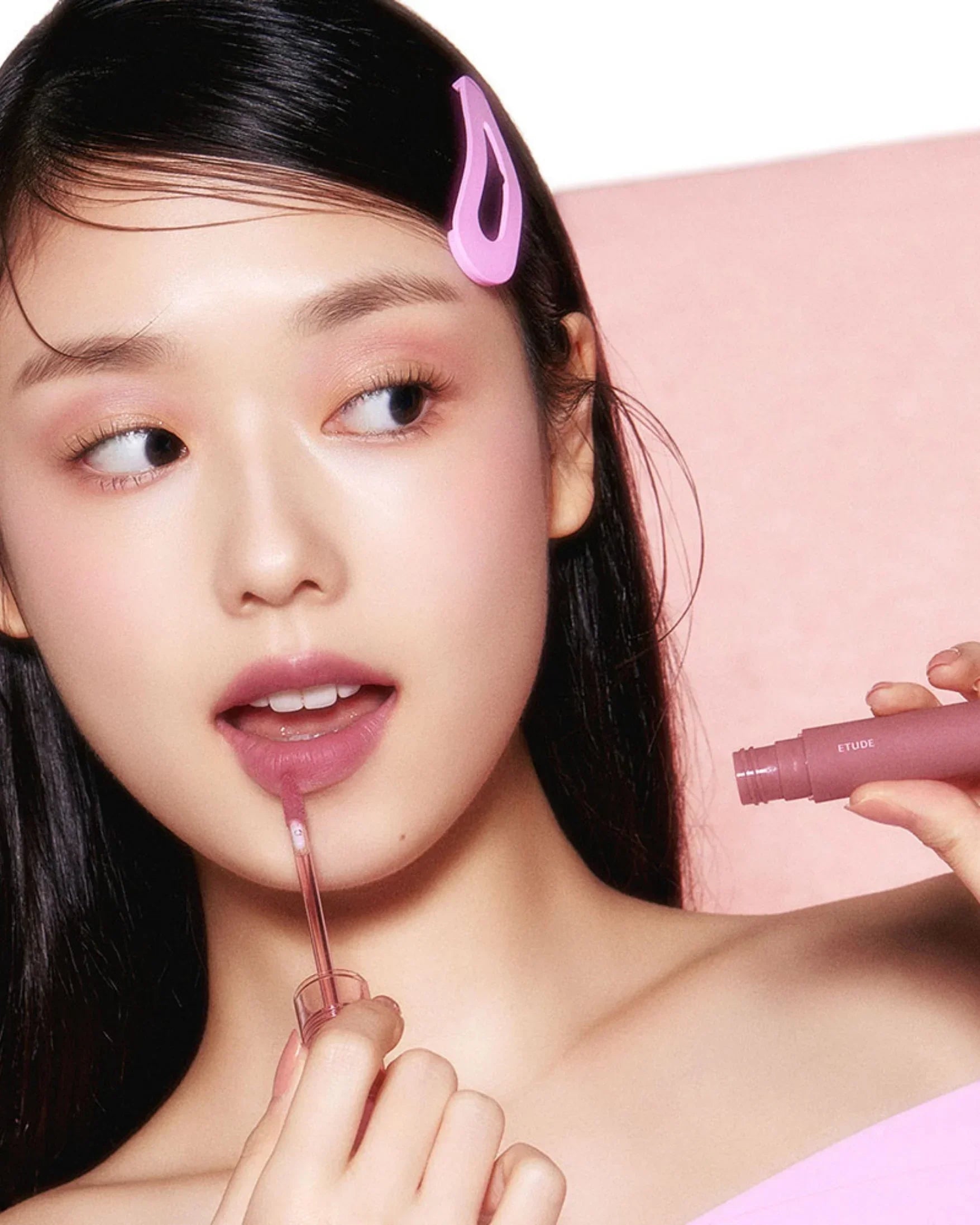Young woman applying pink lip tint with applicator, wearing pink hair clip and off-shoulder top