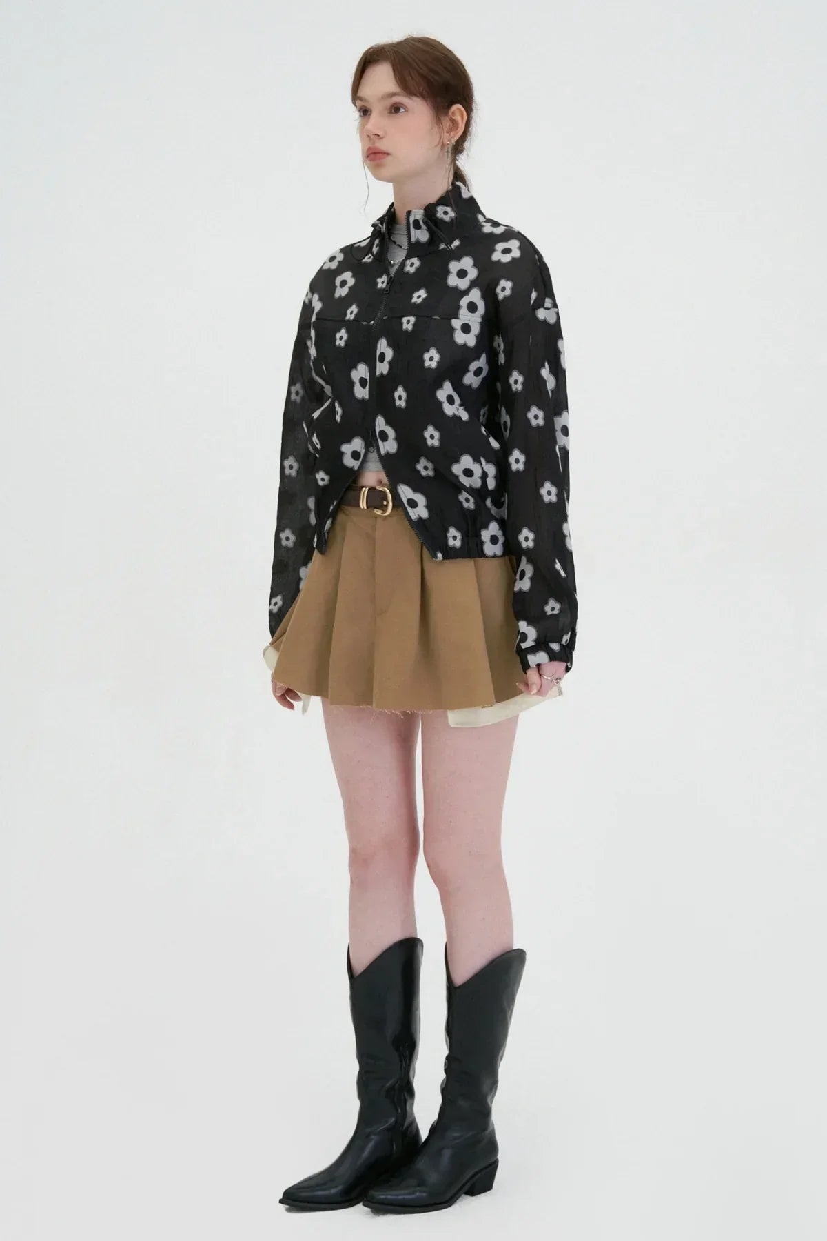 Model wearing black flower print zip-up jacket, beige pleated mini skirt, and black cowboy boots