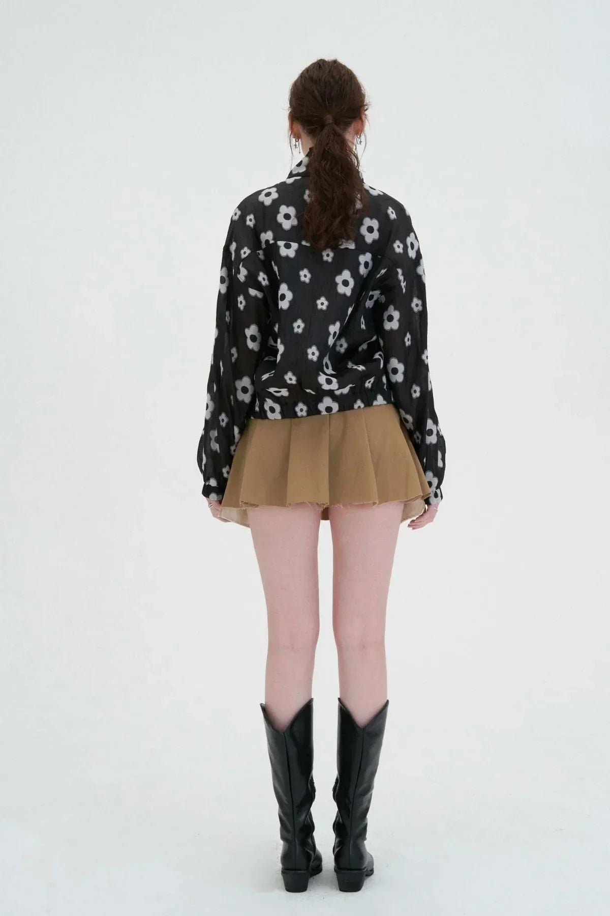 Woman wearing black floral zip-up jacket, brown pleated skirt, and black knee-high boots standing against white background