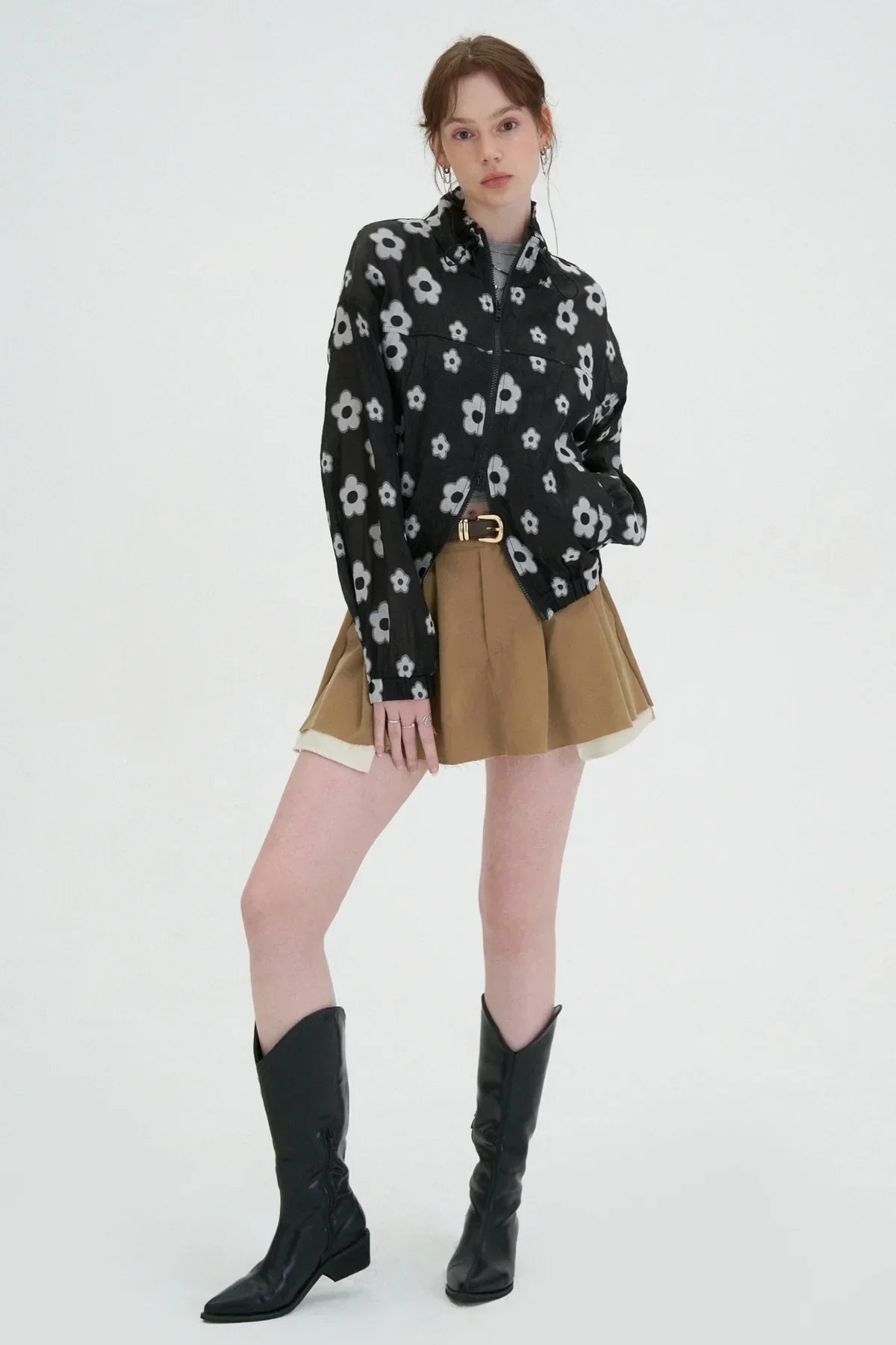 Woman wearing black zip-up jacket with white flower print, tan pleated skirt, and black knee-high boots