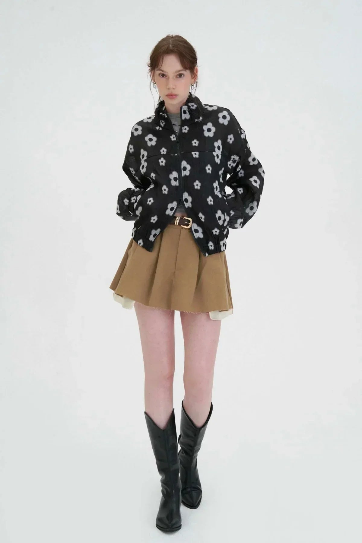 Fashion model wearing black floral zip-up jacket, brown pleated skirt, and black boots in studio