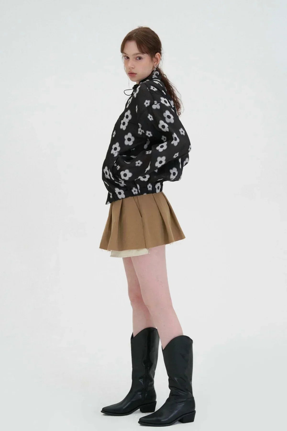 Side view of woman wearing black floral zip-up jacket, brown pleated skirt, and black knee-high boots on white background