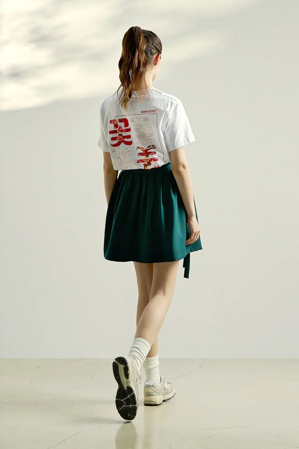 Rear view of woman wearing white graphic t-shirt with red print, dark green skirt, white socks, and sneakers