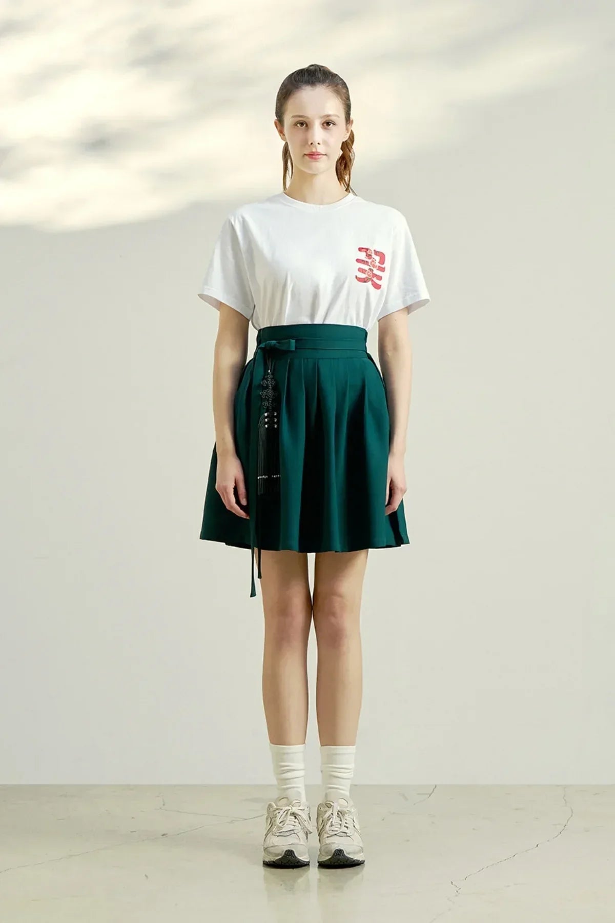 Woman wearing white graphic t-shirt with red design and teal pleated skirt with waist tie