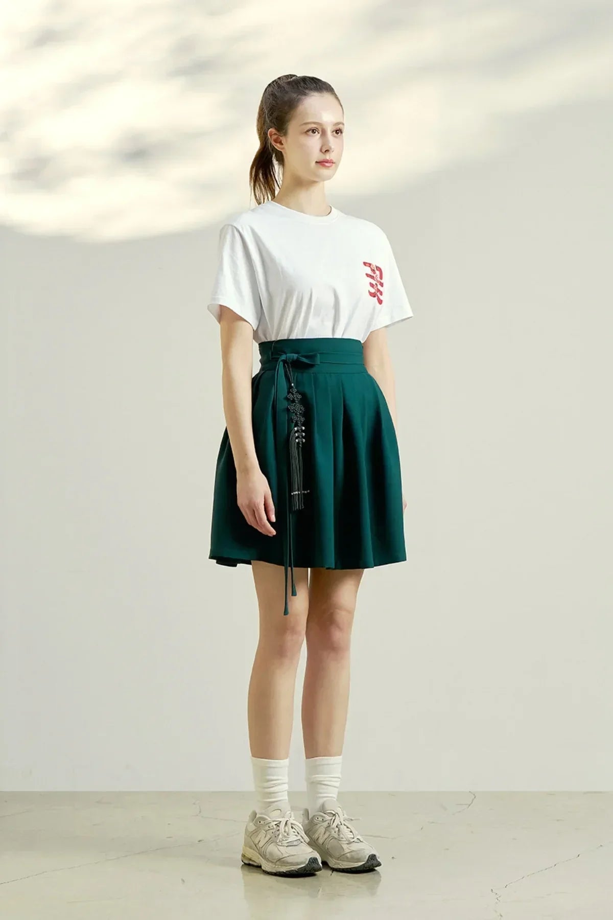 Young woman standing in white graphic t-shirt and dark green pleated skirt with tassel belt, white socks and sneakers