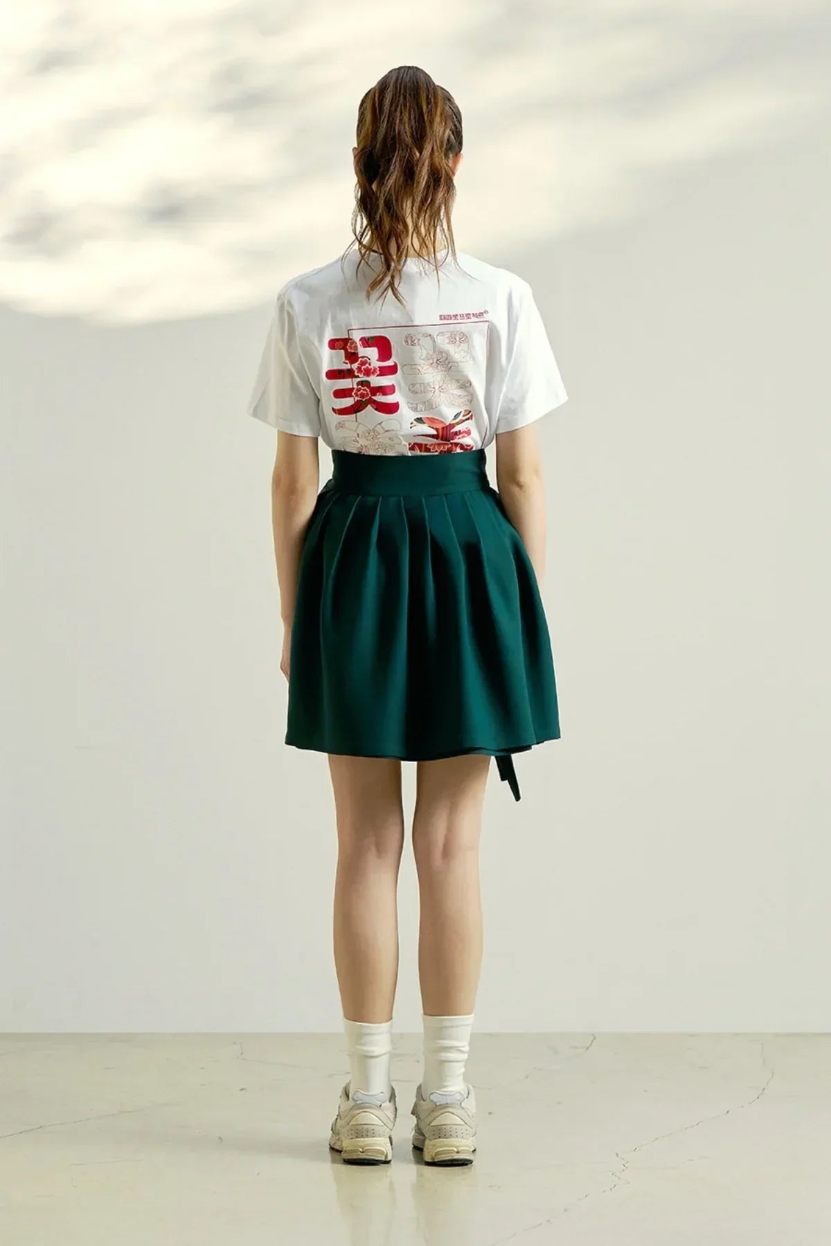 Woman wearing white graphic flower t-shirt and dark green pleated skirt, standing on polished floor