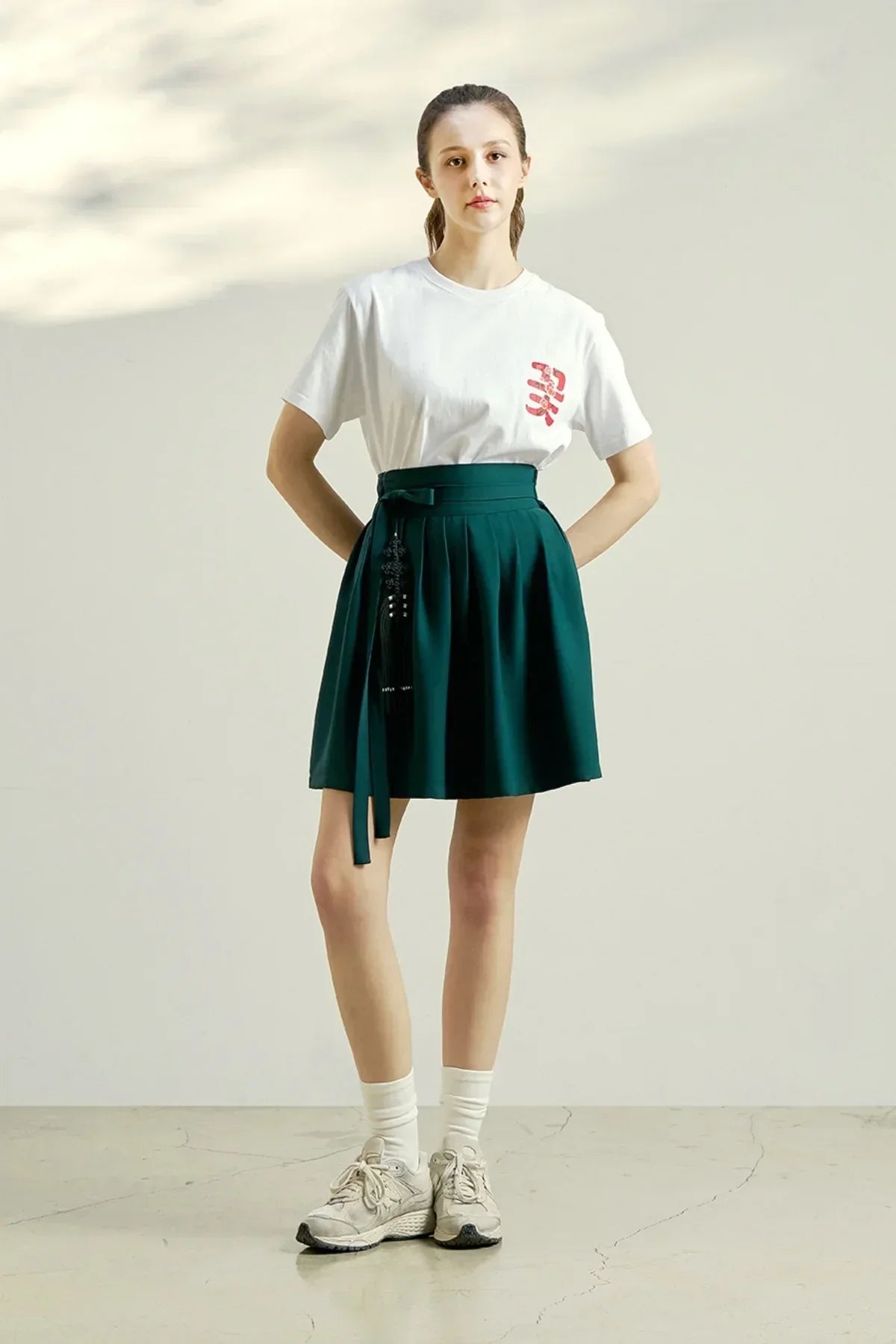 Young woman in white graphic t-shirt and pleated dark green skirt with tassel detail in studio