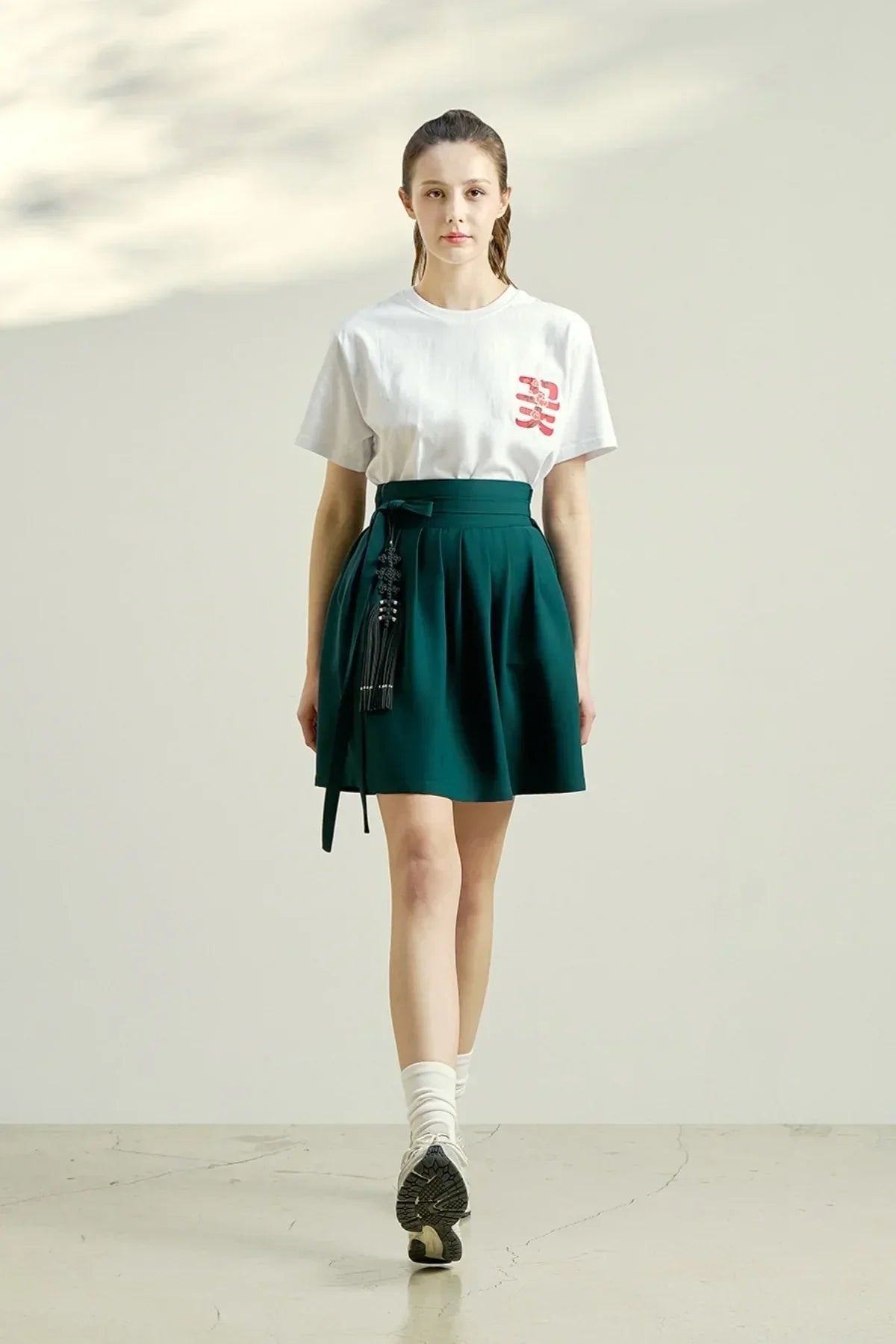 Woman wearing white graphic t-shirt and green pleated skirt with black tassel accessory