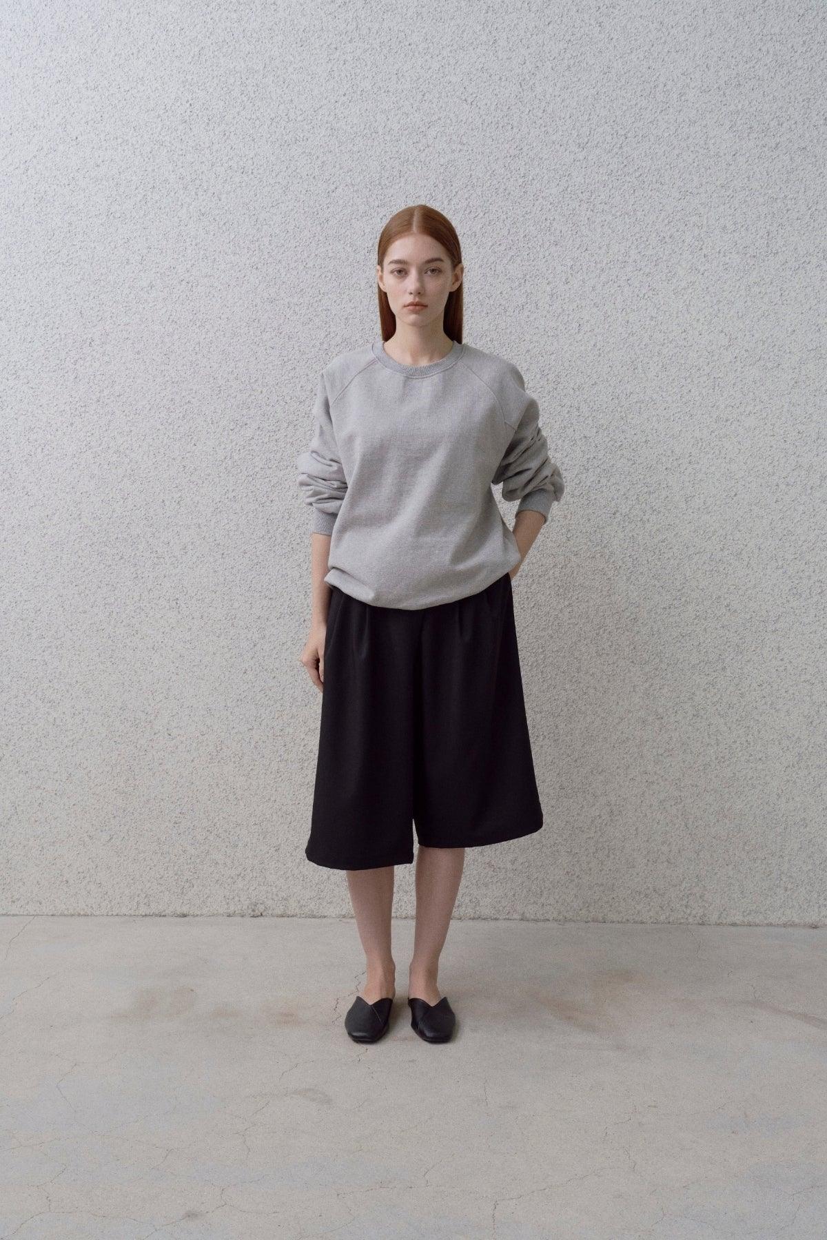 Woman wearing grey round neck sweatshirt, black wide-leg knee-length shorts, and black slip-on shoes against a textured grey wall