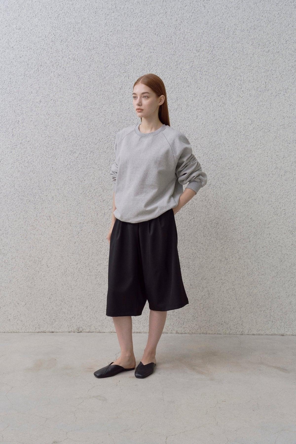 Young woman wearing a grey round neck sweatshirt, black wide shorts, and black slip-on shoes against textured wall