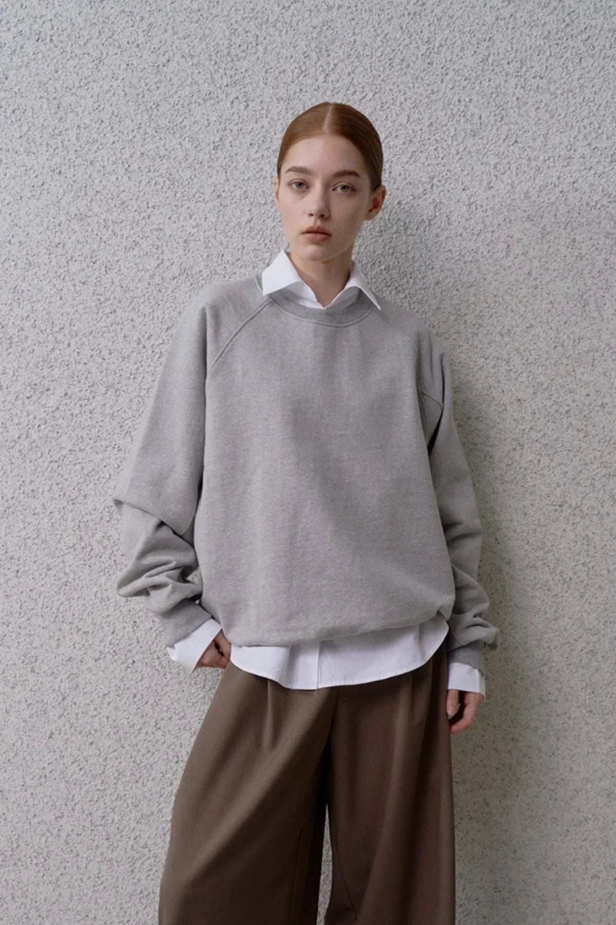 Model wearing grey round neck sweatshirt over white shirt and brown wide-leg pants against grey textured wall