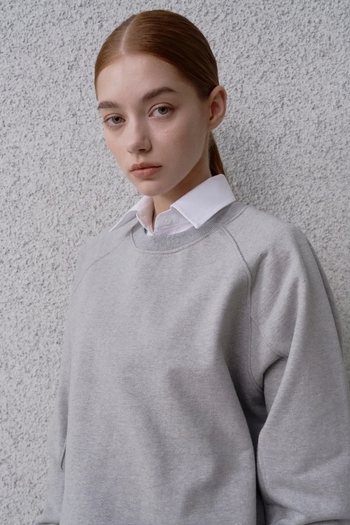 Young woman wearing a grey round neck sweatshirt over a white collared shirt against textured wall