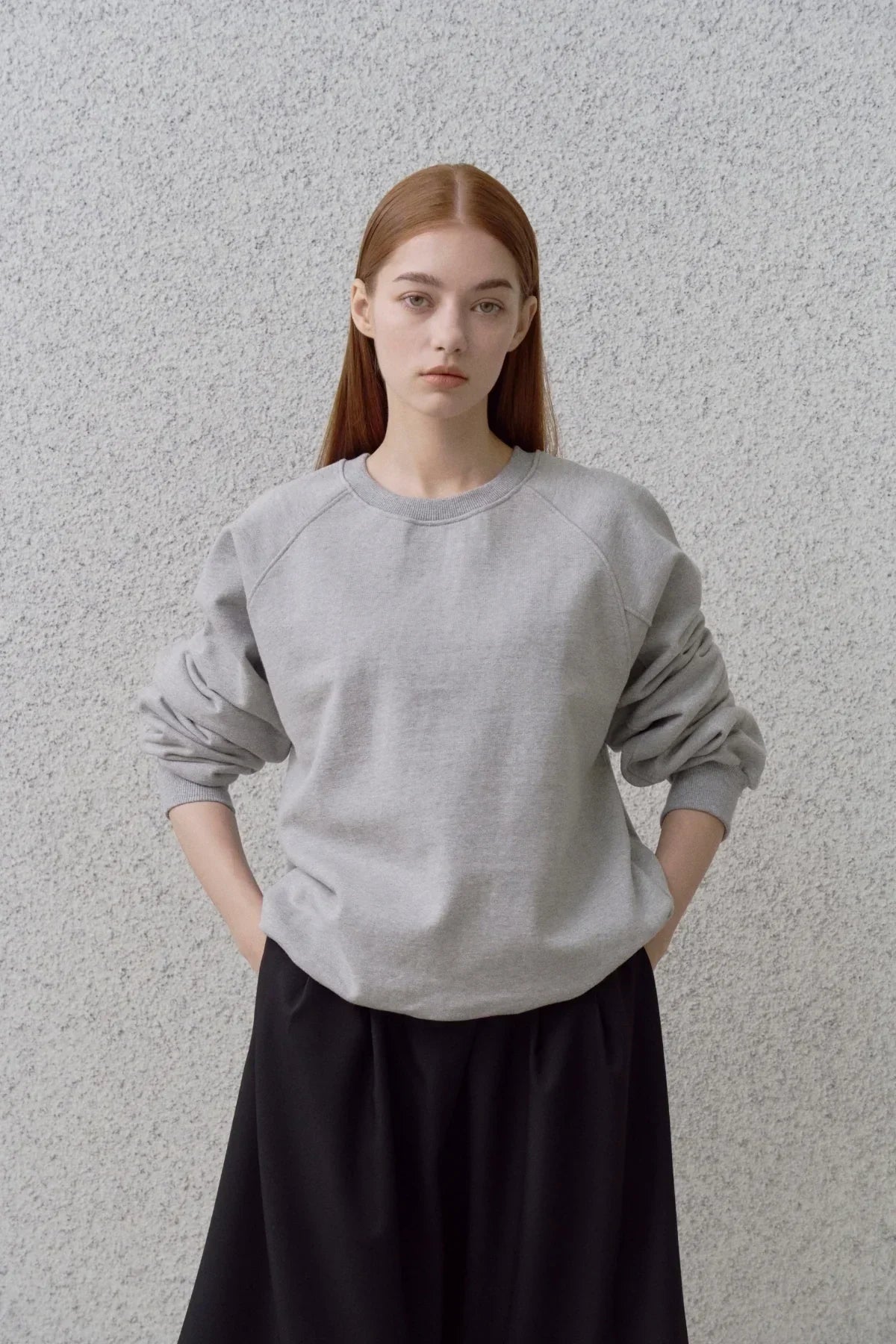 Young woman wearing a grey round neck sweatshirt and black skirt against textured grey wall