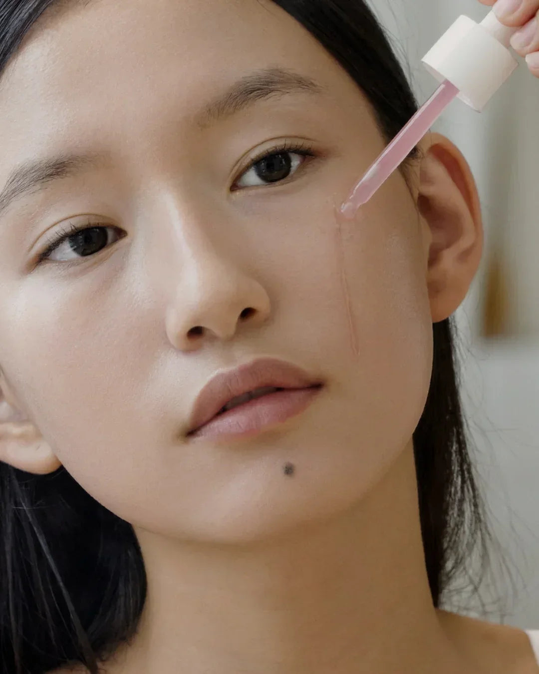 Close-up of woman applying pink serum to cheek with dropper, natural skincare routine