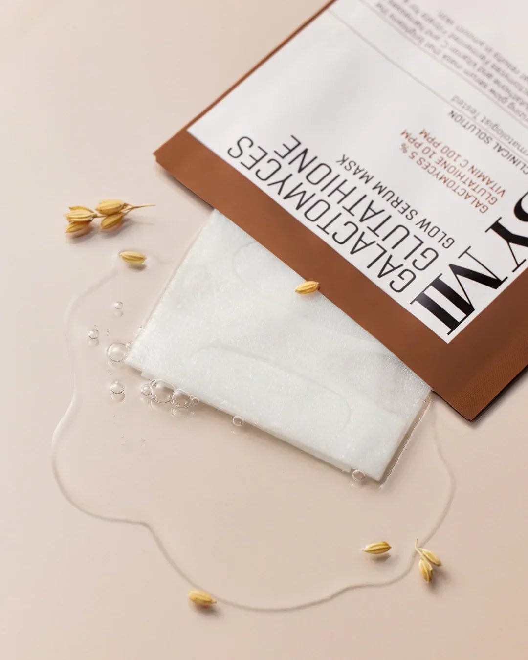 Galactomyces Glutathione Glow Serum Mask partially out of brown and white packaging with serum droplets and grains on light background
