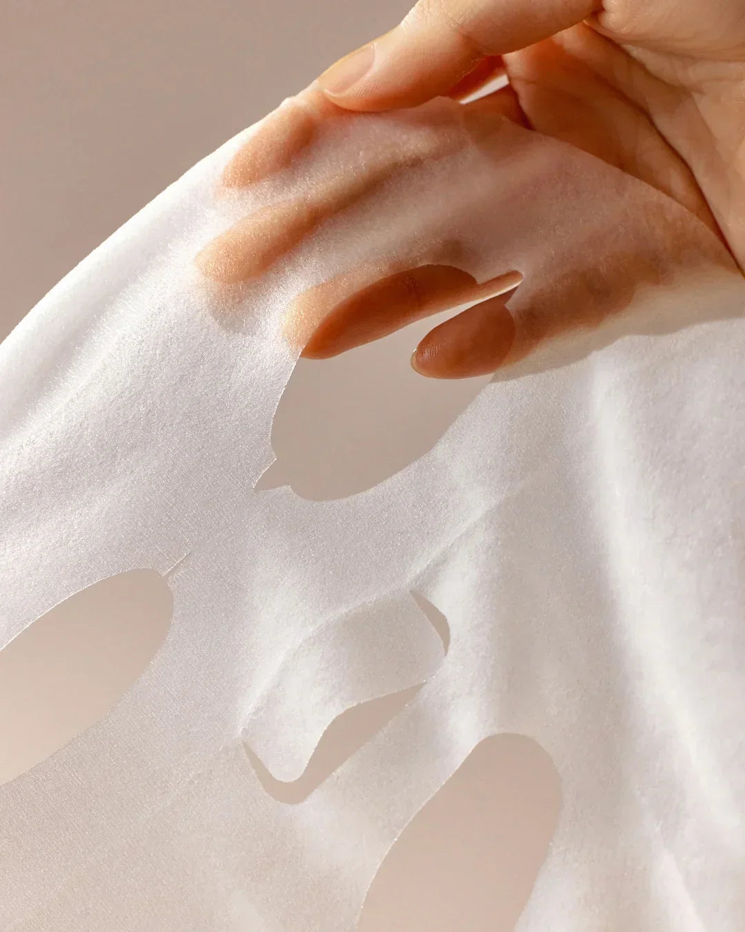 Hand holding a white translucent sheet face mask with cutouts for eyes and mouth