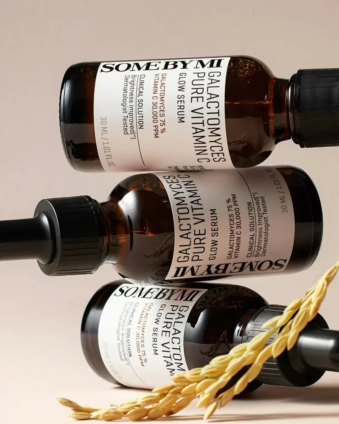 Three amber bottles of SOME BY MI Galactomyces Pure Vitamin C Glow Serum with white labels and a wheat stalk on beige background
