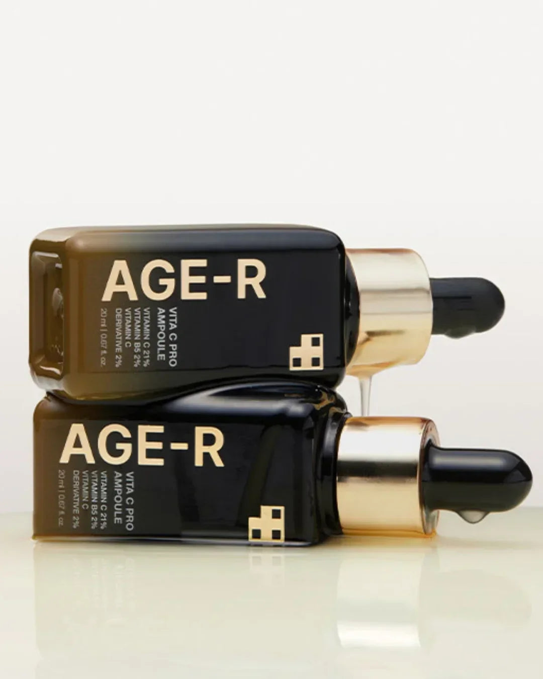 Two black AGE-R Vita C Pro Ampoule skincare bottles with gold droppers stacked on glossy surface
