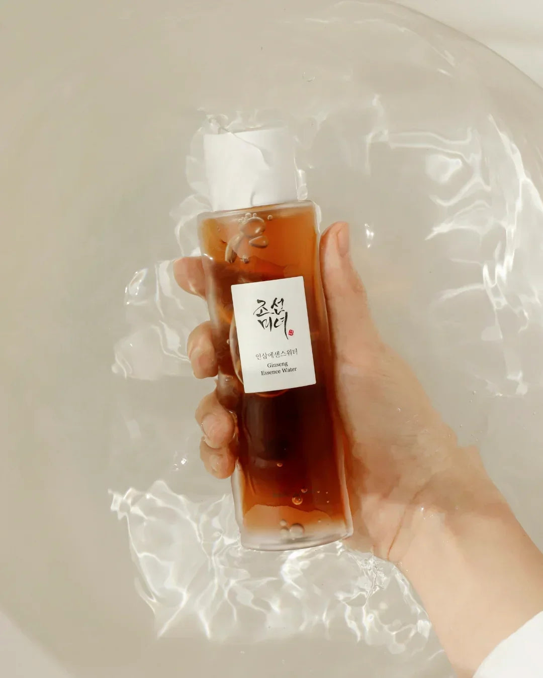 Hand holding a brown bottle of Ginseng Essence Water submerged in clear water with light reflections